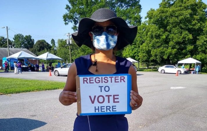 Woman in hat holding Register to Vote sign in parking lot