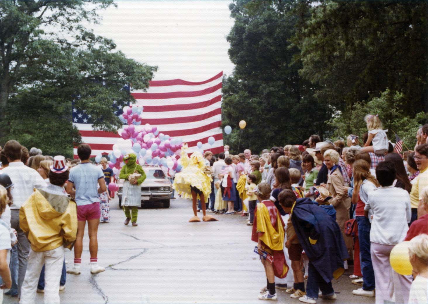 Casually dressed spectators watch a neighborhood Fourth of July parade with a gigantic American flag.