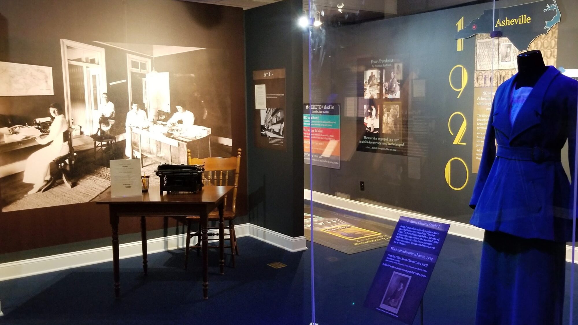 Museum display with blue dress in case in front of wall with picture of four women in white dresses sitting in a suffrage office in 1920